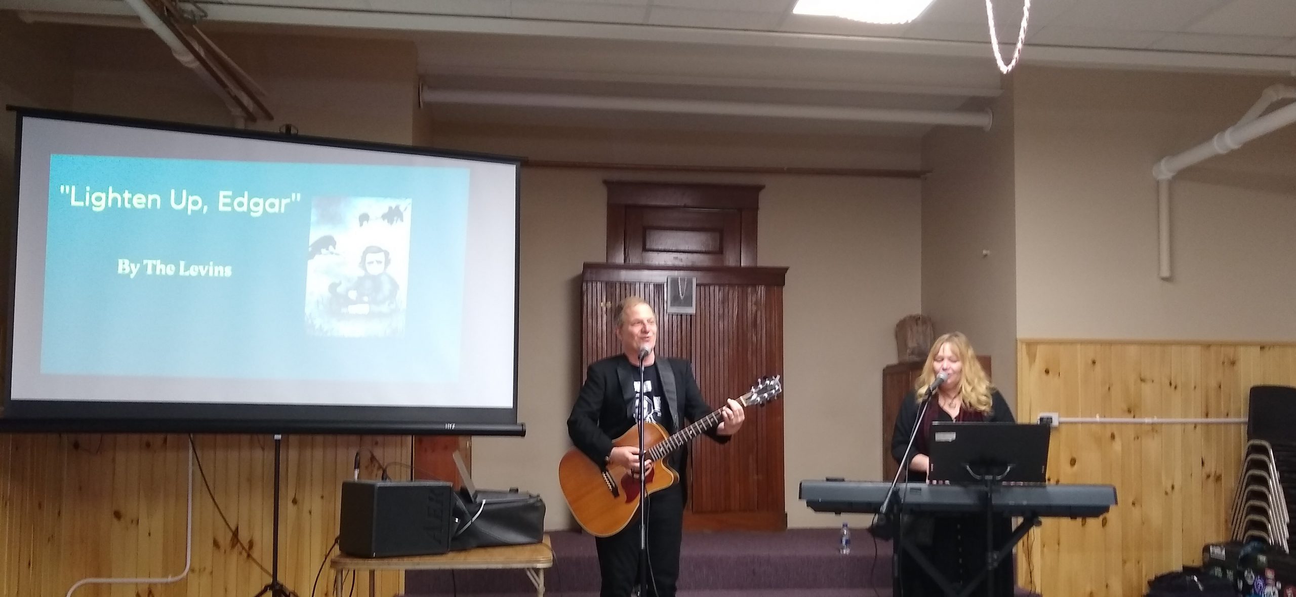 Ira plays the guitar and Julia plays the keyboard as they stand next to a projector screen. The screen says the title of their song, "Lighten Up, Edgar" and there's a picture of Edgar Allan Poe imagined as what he would have looked like as a child