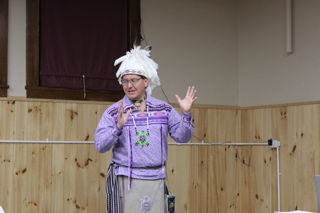 Perry Ground indicates with his hands as he talks. He is wearing the regalia of the Haudenosaunee peoples
